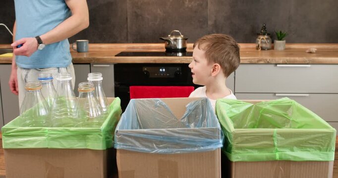 Playful boy drops small plastic piece into blue lined recycling box at home. Child sorts waste with father placing item into container during cleanup task
