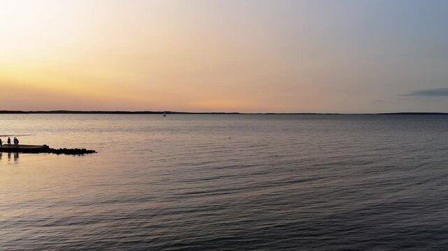 Drone video glides over Hanko, Finland&rsquo;s serene Baltic shore at golden hour as silhouettes linger on the piers, calm water reflecting a soft orange horizon, peaceful dusk, wide panoramic aerial video