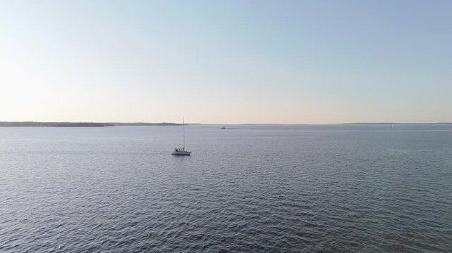 Serene aerial footage of a single sailboat drifting on tranquil open water beneath a pastel sky near Hanko, Finland, with gentle ripples, soft golden hour lighting, and minimal horizon