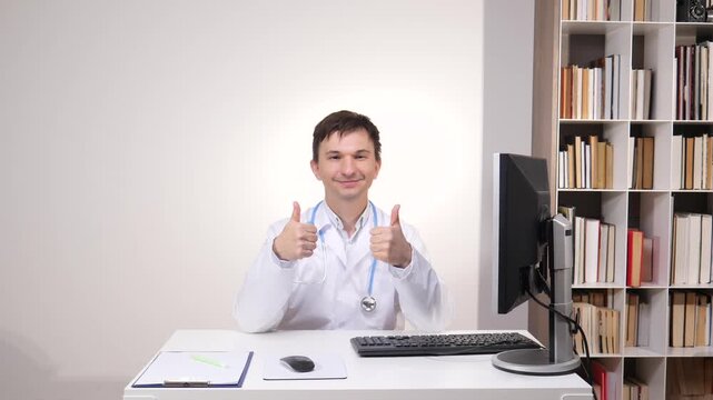 Male doctor in a white lab coat and stethoscope sitting at a desk in his office, giving two thumbs up, expressing approval and satisfaction with medical care