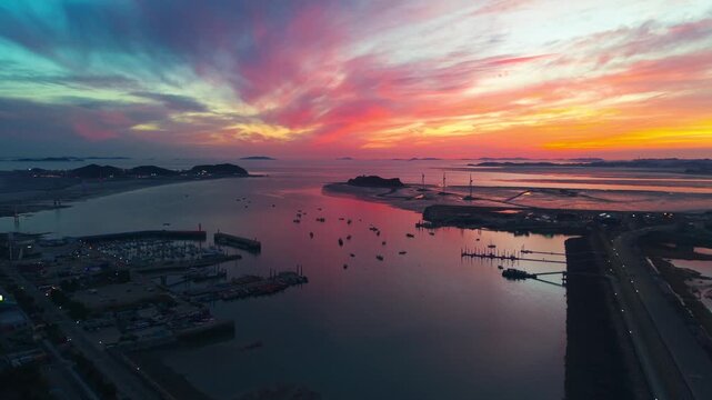 Aerial flyover Jeongok Marina toward silhouetted Nue Island wind turbines and Tando Sea Road at fiery spring sunset with Jebudo and scattered islands reflecting in calm Ansan coastal waters