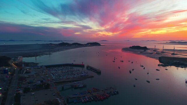 Aerial pull-back pan from Jeongok Marina at vivid spring sunset revealing Tando Sea Road, Nue Island, silhouetted wind turbines and scattered boats reflecting in golden Korean tidal waters