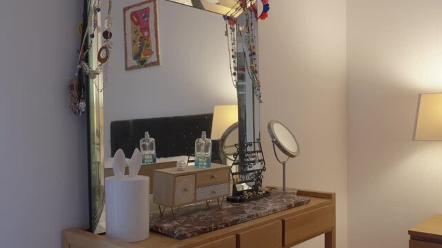 Slow arc shot of a wooden vanity dresser with large mirror, perfume bottles, jewelry organizer, and round vanity mirror in a warmly lit bedroom.