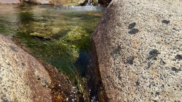 Close-up detail of clear stream water cascading over and around rough, textured granite rocks and smooth river stones, creating ripples and small bubbles.