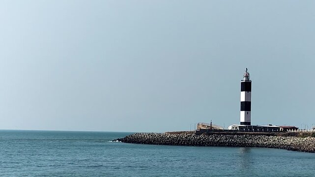 Static view of striped lighthouse on rocky breakwater with calm sea and clear sky. Ideal for maritime themes, navigation concepts, coastal travel visuals, and minimal background shots.