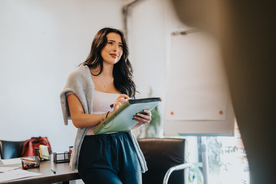 Young woman student holding a clipboard and smiling while preparing notes. A student presents ideas near a flip chart in a relaxed workspace.