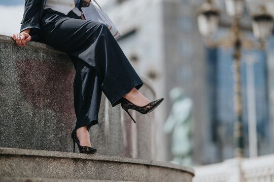 Business person wearing a suit and high heels sits on an urban ledge with crossed legs. A close cropped view rephrases the scene as a stylish professional resting outdoors in the city.