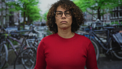 Woman wearing glasses and red sweater stands on street beside parked bicycles with canal in view facing camera  determination. © Krakenimages.com