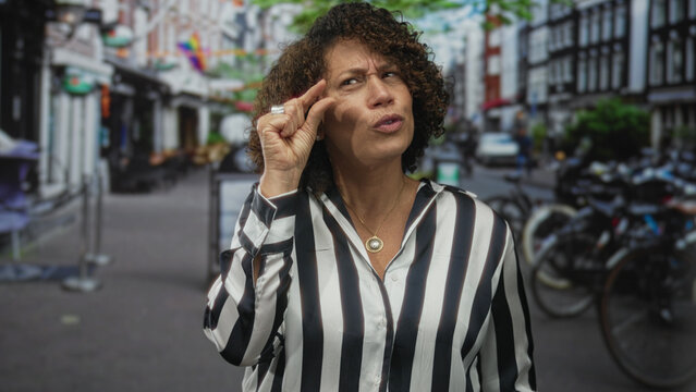 Woman pinches fingers near her face on a busy urban street with bicycles and brick buildings in view; skepticism.