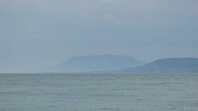 Amazing view of the famous Badacsony Mountain on a cloudy and humid autumn morning. Blue wavy water of Lake Balaton in the foreground. Scenic landscape from Hungary with a different shades of blue.