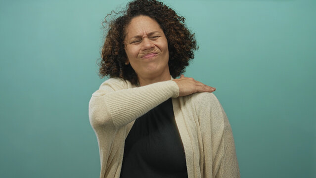 Hispanic woman clutching her left shoulder with a wince in pain against a teal studio wall; discomfort.