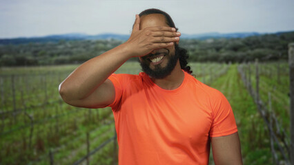 Naklejka premium Man covers eyes with hand and smiles in forest vineyard rows wearing bright orange shirt and beard; playful shyness.