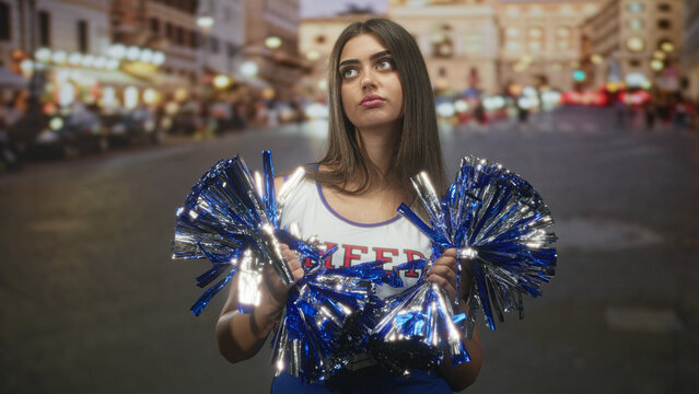 Woman cheerleader holding blue metallic pompoms with both hands, gazing upward while standing in a city street lined with lights at dusk; contemplation.