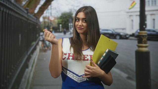 Young woman cheerleader in blue and white uniform holding a yellow clipboard and books, beckons with finger and smiles on a street beside iron railings; confidence youth.