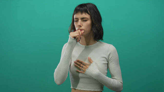 Young hispanic woman coughs with hand on chest and eyes closed against green backdrop in studio; concern.