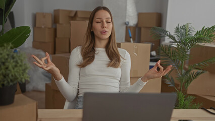 Fototapeta premium Woman seated at laptop amid stacked moving cardboard boxes in a new home building, eyes closed with hands in meditation mudra; calm focus.