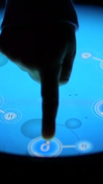 Close up shot of a finger using an interactive multimedia display, tapping on icons representing chemical elements like hydrogen and oxygen to create a water molecule in a scientific simulation