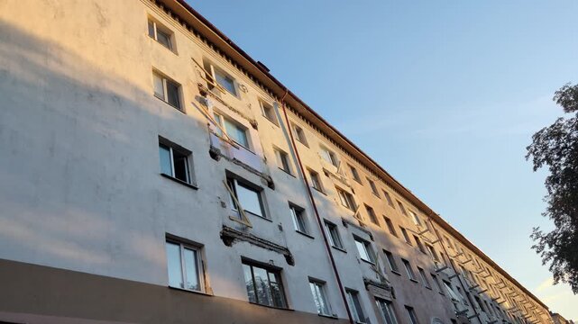 Exterior view of multi-story residential building undergoing major renovation. Numerous balconies are demolished or partially removed, revealing concrete structure. Sunlight illuminates facade against
