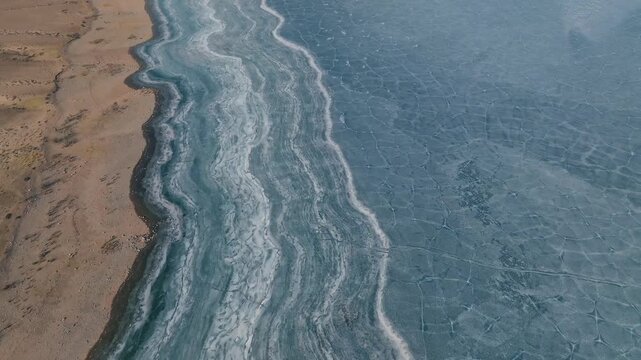 Drone footage of the Orto-Tokoy Reservoir shoreline with layered frozen waves and cracked blue ice patterns beside a sandy bank in Issyk-Kul Region, Kyrgyzstan
