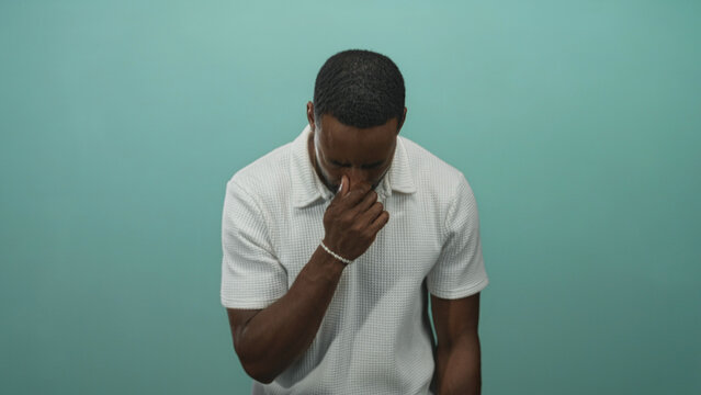 Young african american man coughing into hand with head bowed, wearing white textured polo shirt in teal studio with teal background and soft light; concern.