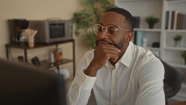 Man wearing white shirt and round glasses rests chin on hand at office desk while studying a monitor; pensive concentration.