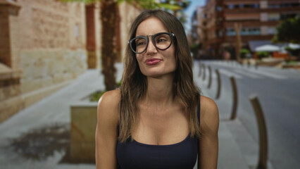 Naklejka premium Woman wearing glasses and tank top points fingers upward on city street; celebration excitement spontaneity.