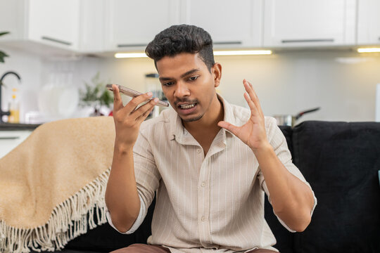 Indian man at home records voice message on smartphone speaks clear checks mic level before reply. Arabian guy on sofa talks in audio chat uses gesture for emphasis sends message and ends recording