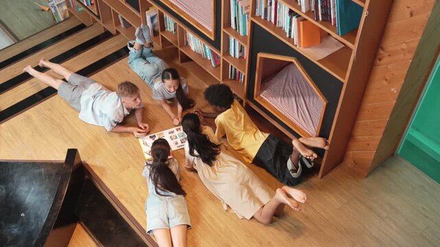 Group of diverse children lying down in circle while reading a book at library. Top view of girl sharing a magazine while pointing at interested topic and talking with lovely friends. Edification