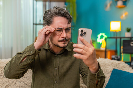 Middle-aged man at home in glasses squints reading tiny smartphone text worries about poor eyesight. Young adult guy on sofa raises phone closer tests vision clarity blinking to focus on screen words