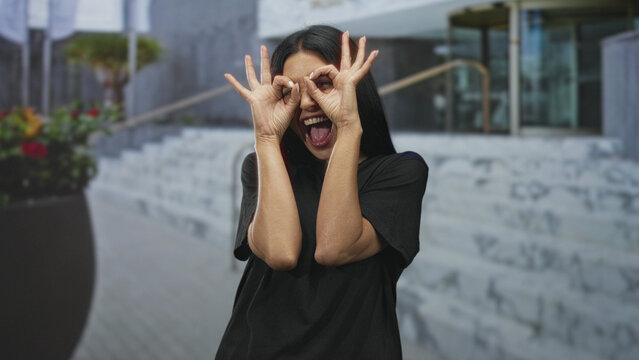 Woman making hand binoculars to eyes at building entrance, hands framing face with tongue out and laughing wide smile; playful spontaneity.
