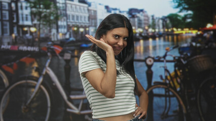 Naklejka premium Woman in striped crop top waves hand by face and poses beside bicycle in studio canal scene with city lights and bridge railing; playful coyness.
