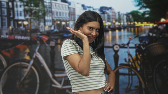 Woman in striped crop top waves hand by face and poses beside bicycle in studio canal scene with city lights and bridge railing; playful coyness.