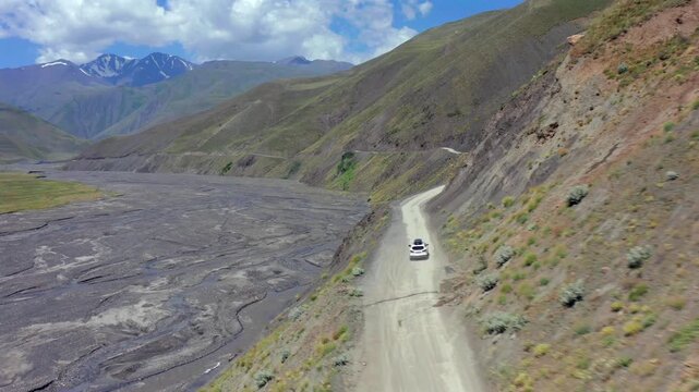 Aerial drone shot of SUV driving along narrow road beside dry river valley in Azerbaijan, showing rugged terrain, erosion patterns and remote mountain travel landscape