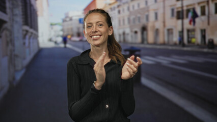 Naklejka premium Woman clapping hands on a sunlit city street, smiling and leaning forward with hair in a ponytail and visible pearl earrings; joy celebration warmth.