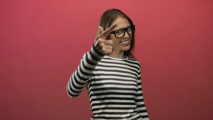 Naklejka premium Woman wearing glasses and striped shirt smiles and raises fists in pink studio; joy playful celebration.