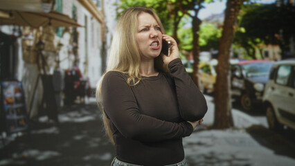 Obraz premium Woman holding smartphone to ear, grimacing and clenching fist on city street; frustration confrontation.