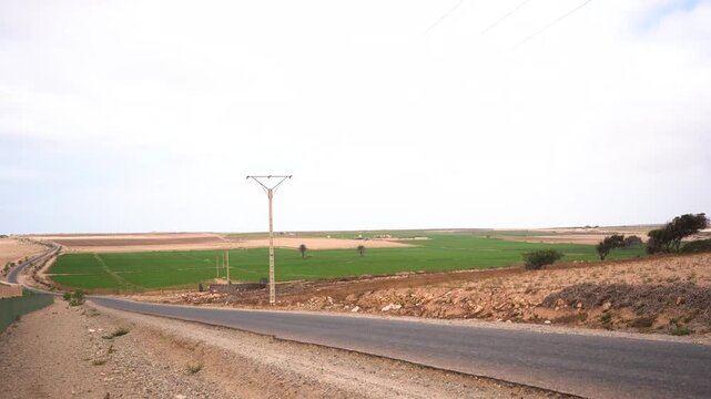 A wide, elevated static shot looking down a paved country road in Morocco under a soft, overcast sky. The landscape features a striking visual contrast, showcasing vibrant green farming North Africa