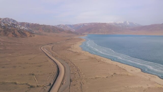 Drone footage of the Orto-Tokoy Reservoir with turquoise water, sandy shoreline, and winding road, framed by snowcapped mountains in Issyk-Kul Region, Kyrgyzstan