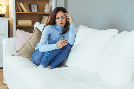 Pensive Young Woman Sitting on White Sofa Looking at Phone in Cozy Living Room