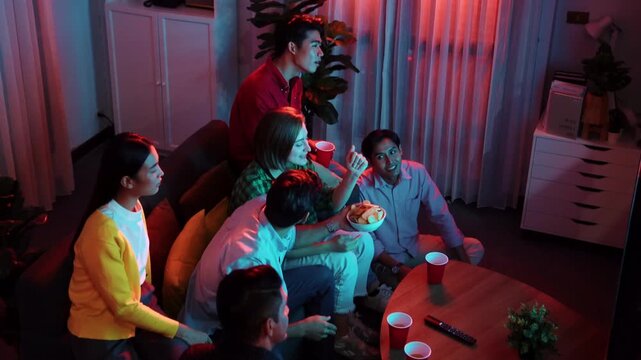Friend cheering football together and clinking glass at living room. Diverse worker or colleague celebrate winning team while clinking glass and drinking alcohol. Woman clapping hand. Convocation.