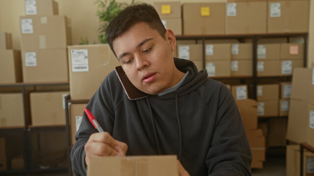 Man holding smartphone to ear writes parcel with red pen using right hand in a building office storage area; focus efficiency.