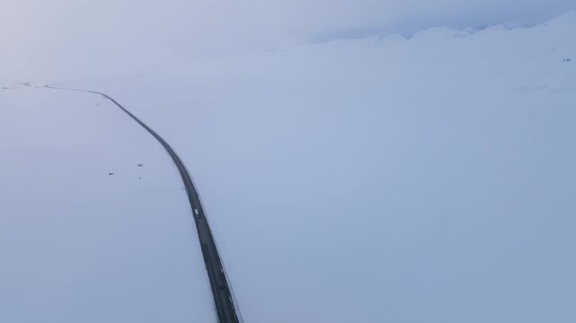 Drone view of a long asphalt road cutting through vast white snowfields at Ala-Bel Pass in the Tien Shan mountains, Kyrgyzstan, with cars driving in winter weather