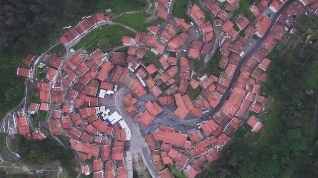 Aerial view of the red-tiled rooftops and winding streets of the coastal village of Cudillero nestled among lush green hills in Cudillero, Asturias, Spain.
