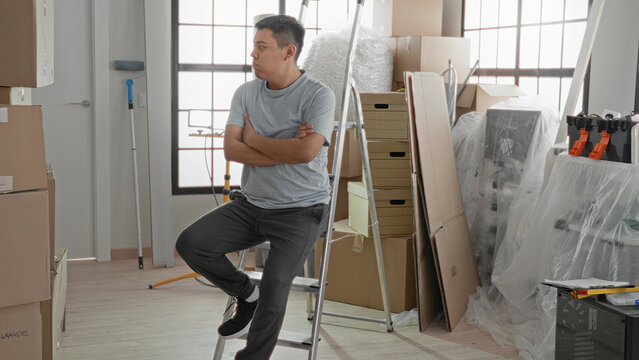 Man sitting on ladder touching chin amid stacked cardboard boxes, packing tape and dolly in a building new home; moving transition uncertainty.