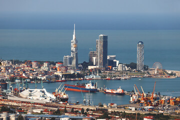 Fototapeta premium High-angle panoramic view of Batumi waterfront, featuring modern skyscrapers like the Alphabetic Tower and Batumi Tower, the industrial sea port with cargo ships, and the Black Sea coast 