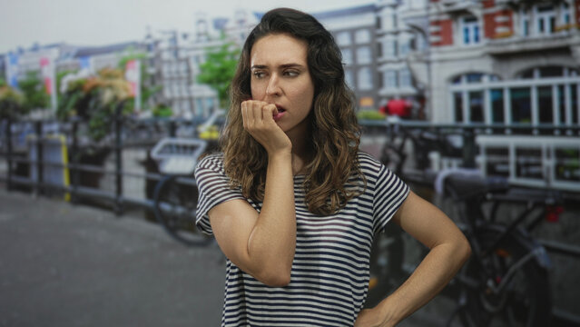 Woman biting her fingernails with hand to mouth by historic urban canal on cobblestone street; anxiety.