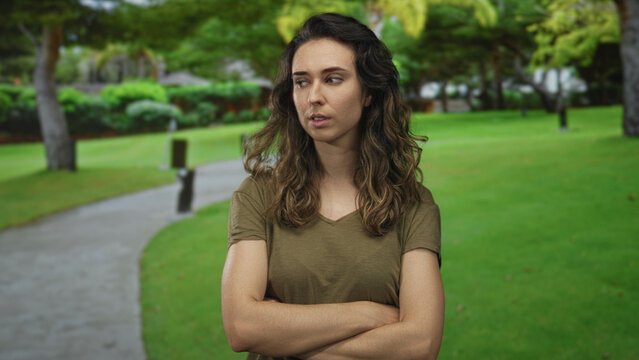 Woman in olive shirt crosses arms and looks sideways in a green park with a paved path under shaded trees; rejection.