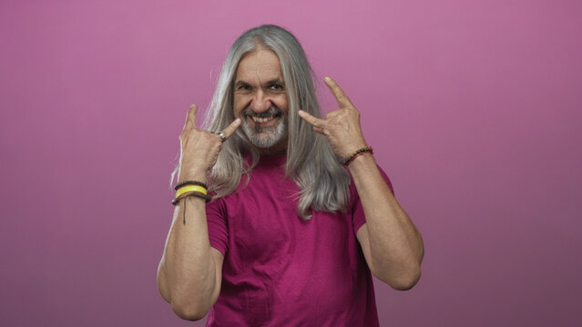Man middle age with hoary long hair grinning while showing rock horns hand gesture in pink studio with bracelets and rings visible; rebellion attitude energy.