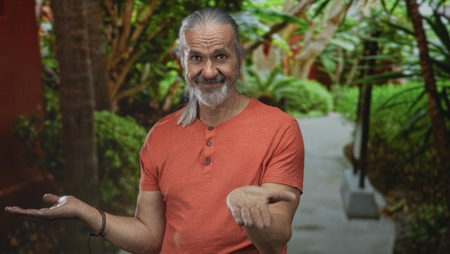 Man with long gray hair and beard holds both palms up on a leafy park path in forest, smiling gently; serenity.
