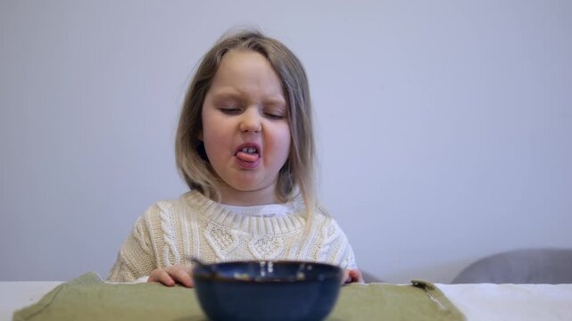 Little girl sitting at the dinner table pushing away a bowl of porridge, making a disgusted face and refusing to eat her meal. Fussy eating habits and picky childhood nutrition concepts